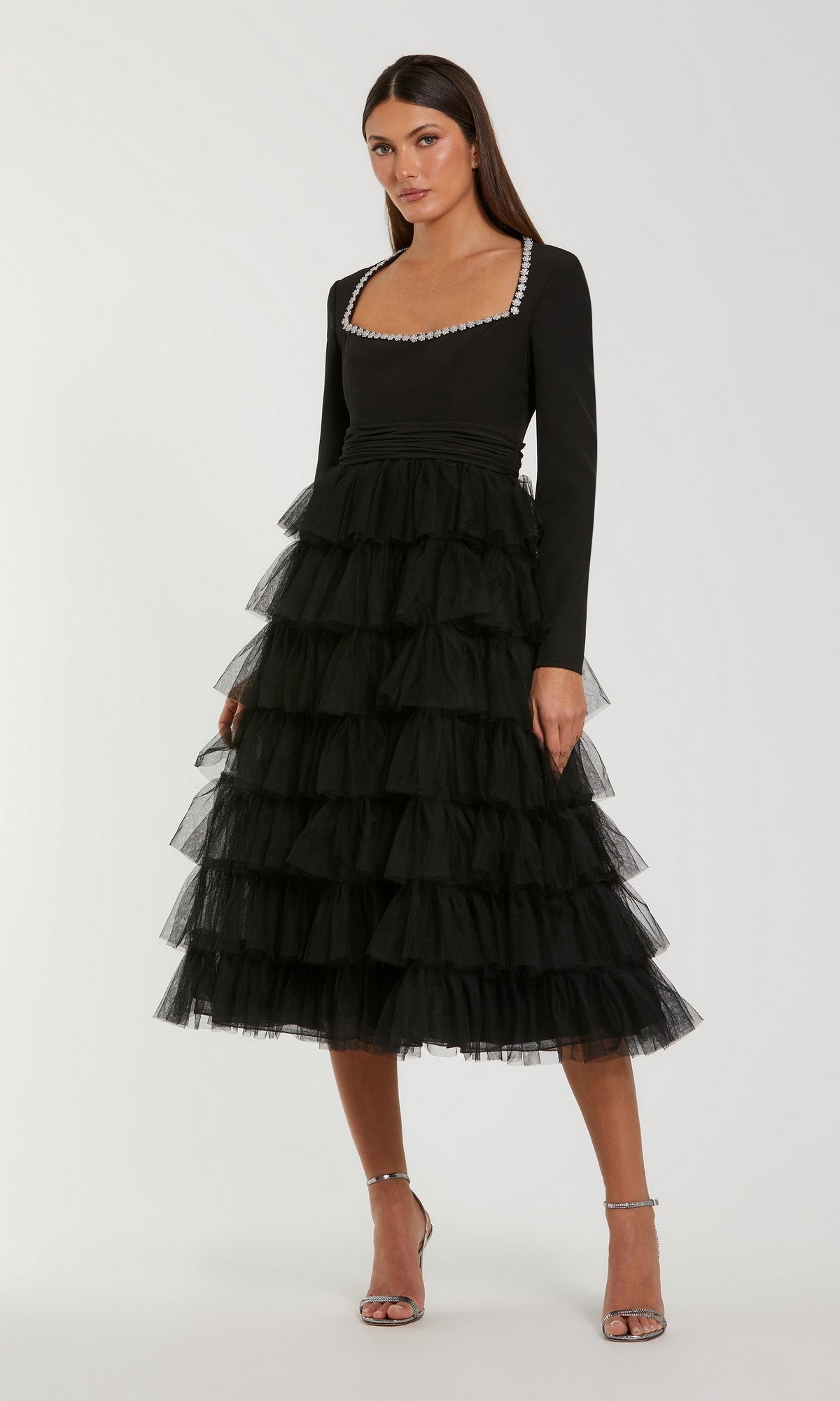 A woman stands against a plain background wearing the Midi Wedding Guest Dress: Mac Duggal 27707 in black, featuring a ruffled tulle skirt, long sleeves, and a pearl-trimmed square neckline, paired with silver high-heeled sandals. This elegant and modest dress offers timeless sophistication for wedding guests.